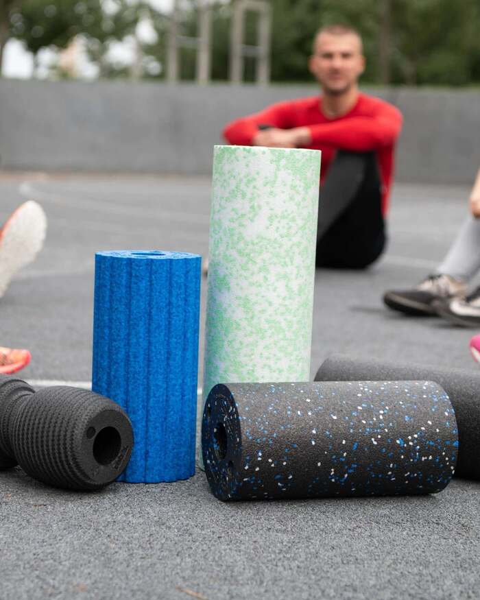 BLACKROLL, Vier Faszienrollen auf Asphalt, dahinter eine Trainingsgruppe im Freien
