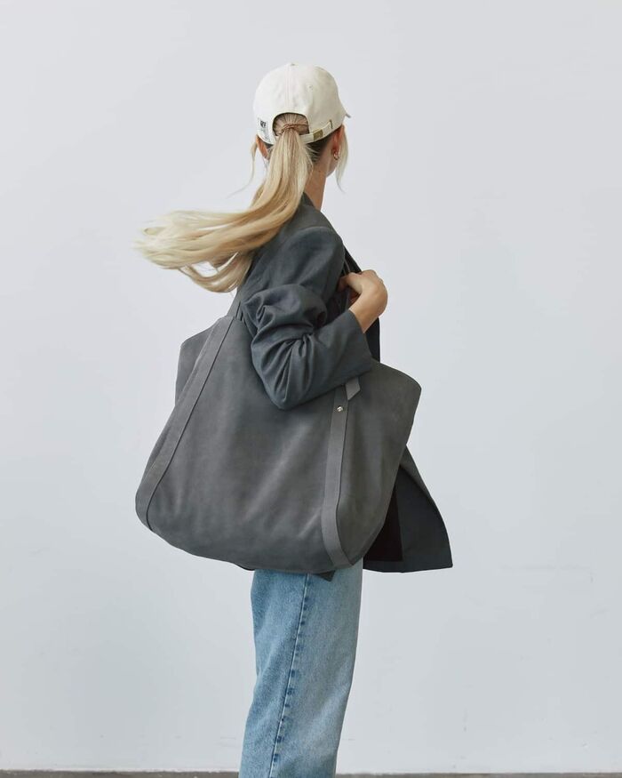 Hey Marly, Frau mit Cap und großer Schultertasche von hinten vor heller Wand