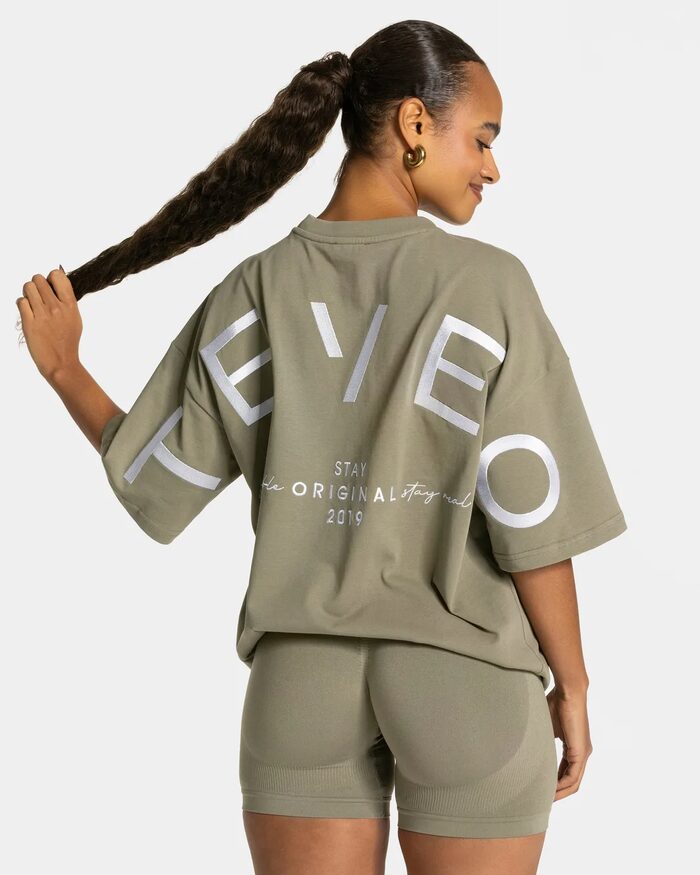 TEVEO, Frau von hinten in khakifarbenem Shirt mit TEVEO-Schriftzug, Zopf in der Hand