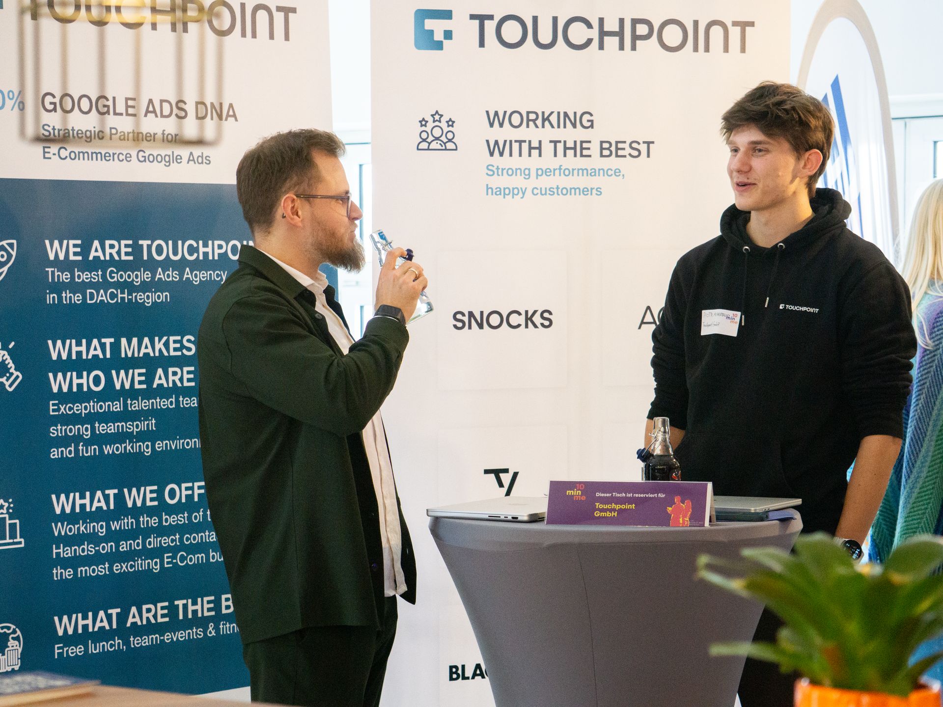 Zwei Kollegen im Gespräch am Touchpoint-Stand vor der Claim-Wand mit Kundenlogos.
